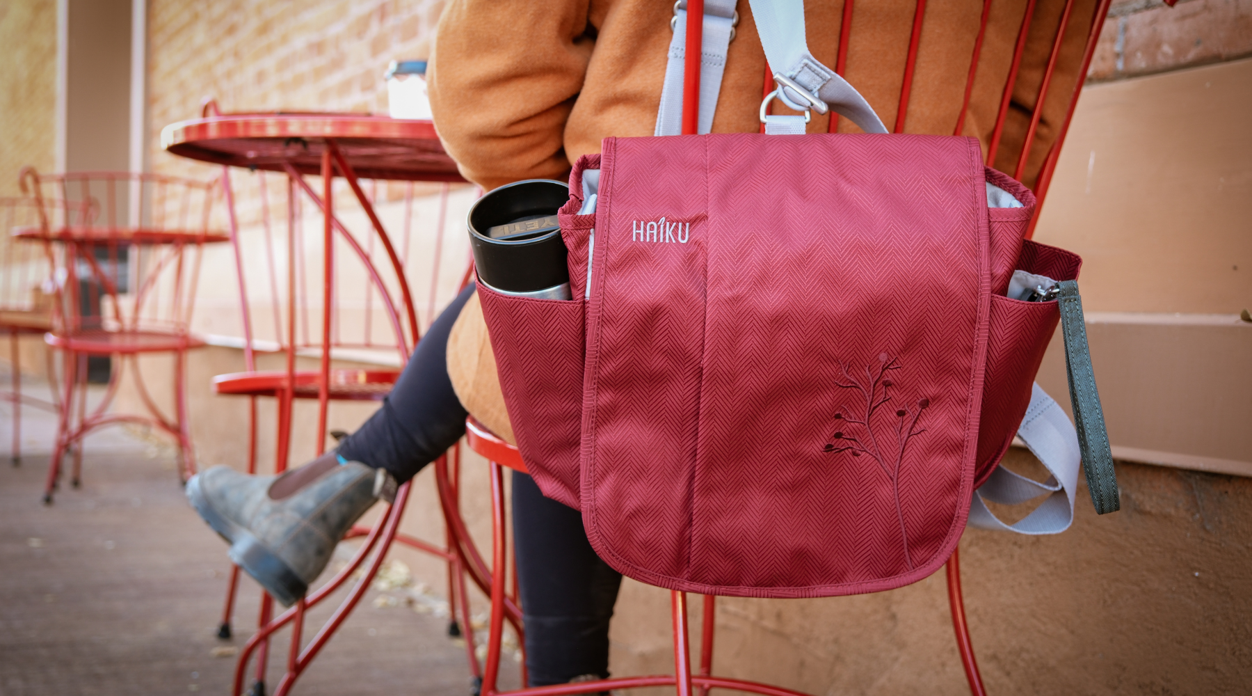 Person sitting at a red outdoor table with a red bag and cup, wearing a orange jacket.